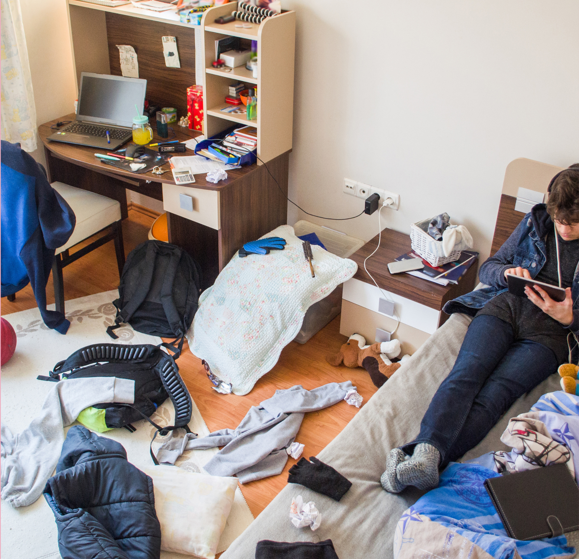Teen sitting on a bed using a tablet in a messy bedroom with clothes and belongings scattered on the floor