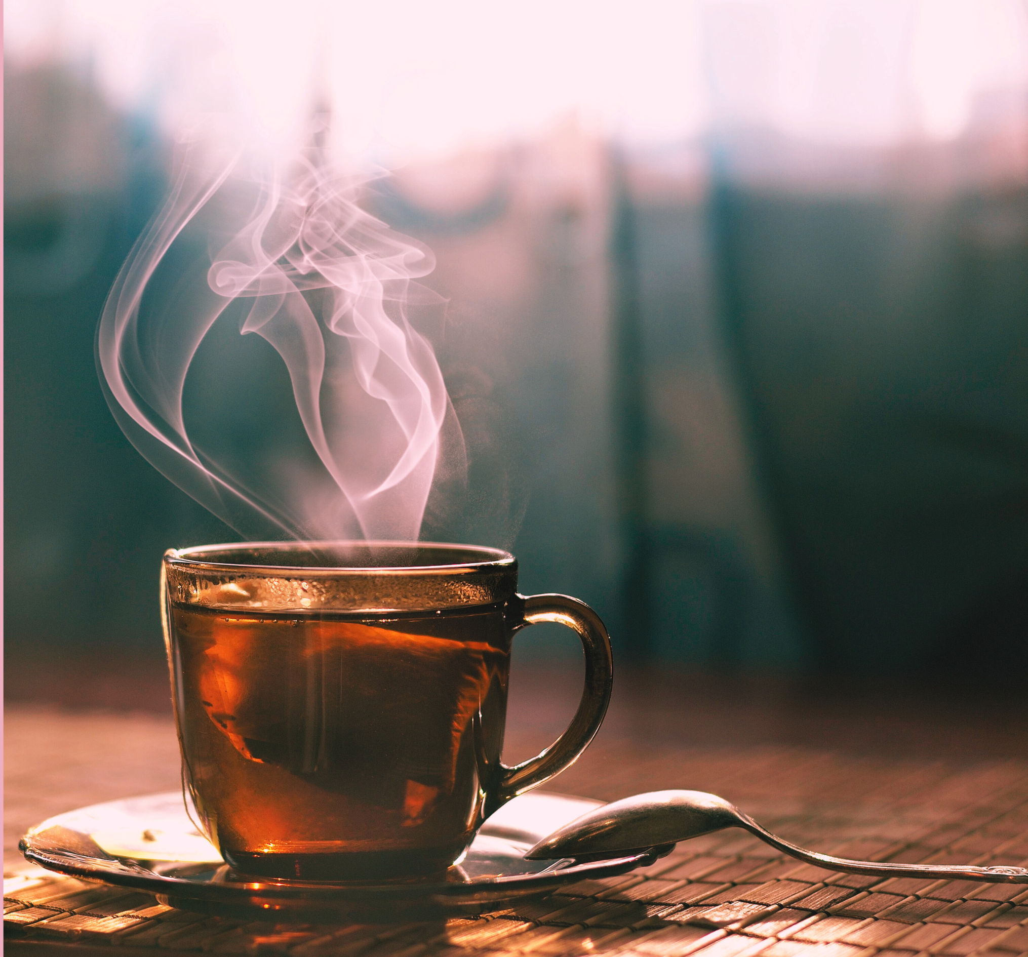 Clear glass cup of tea with steam rising on a wooden surface, with a spoon beside it